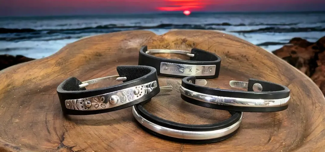 Quatre bracelets en cuir noir et argent massif 925 avec des gravures ethniques et posés sur un plateau en bois avec un coucher de soleil en fond